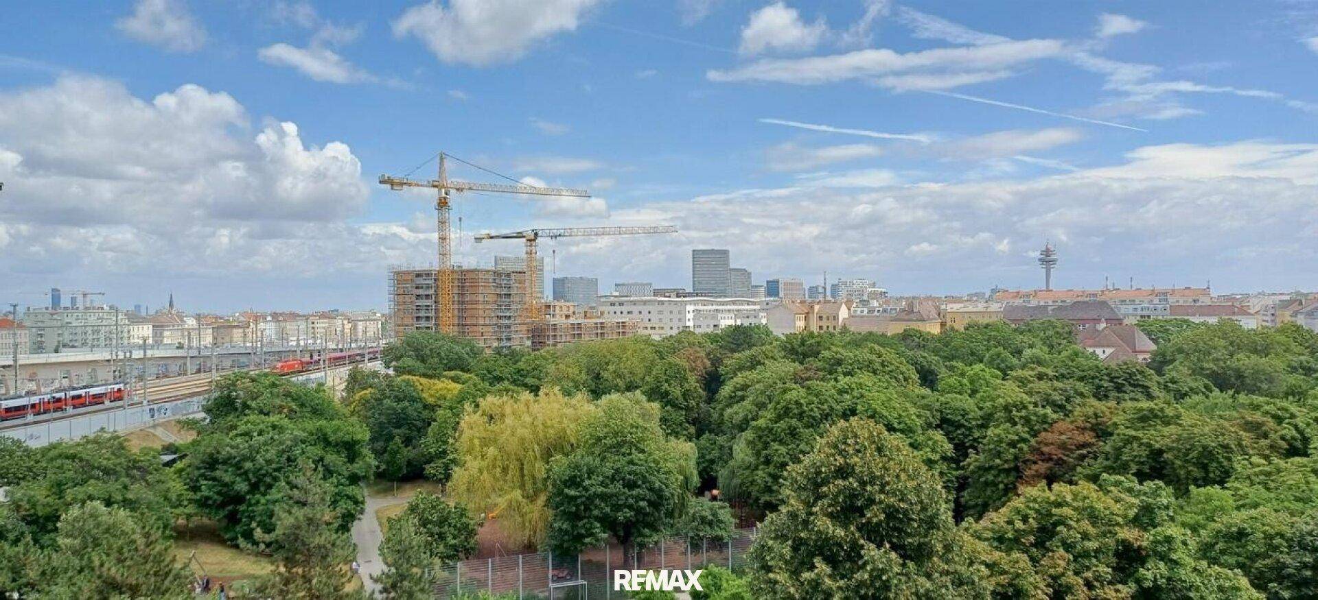 Weitläufiger Blick über die Stadt mit Baustellen, viel Grün und Bahngleisen unter blauem Himmel.