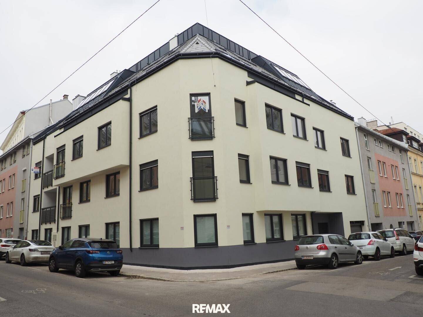 Modernes Eckgebäude mit heller Fassade und dunklen Fensterrahmen an einer Stadtstraße.