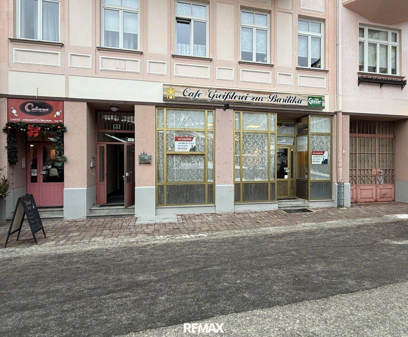 Fassade eines Geschäftsgebäudes mit einem Café-Schild und einem "Zu Kaufen"-Hinweis im Schaufenster.