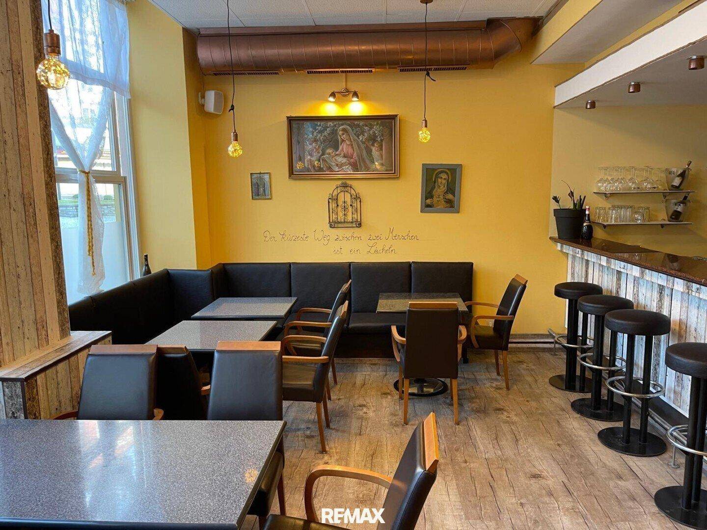 Gastraum mit dunklen Sitzbänken, Holztischen und einer Bar mit Hockern, beleuchtet durch ein großes Fenster.