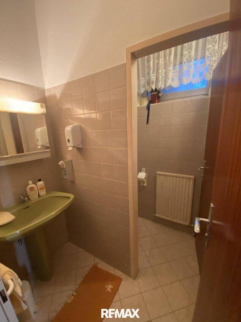 Badezimmer mit Waschbecken, Spiegel und Blick in einen angrenzenden Toilettenbereich, gefliest.