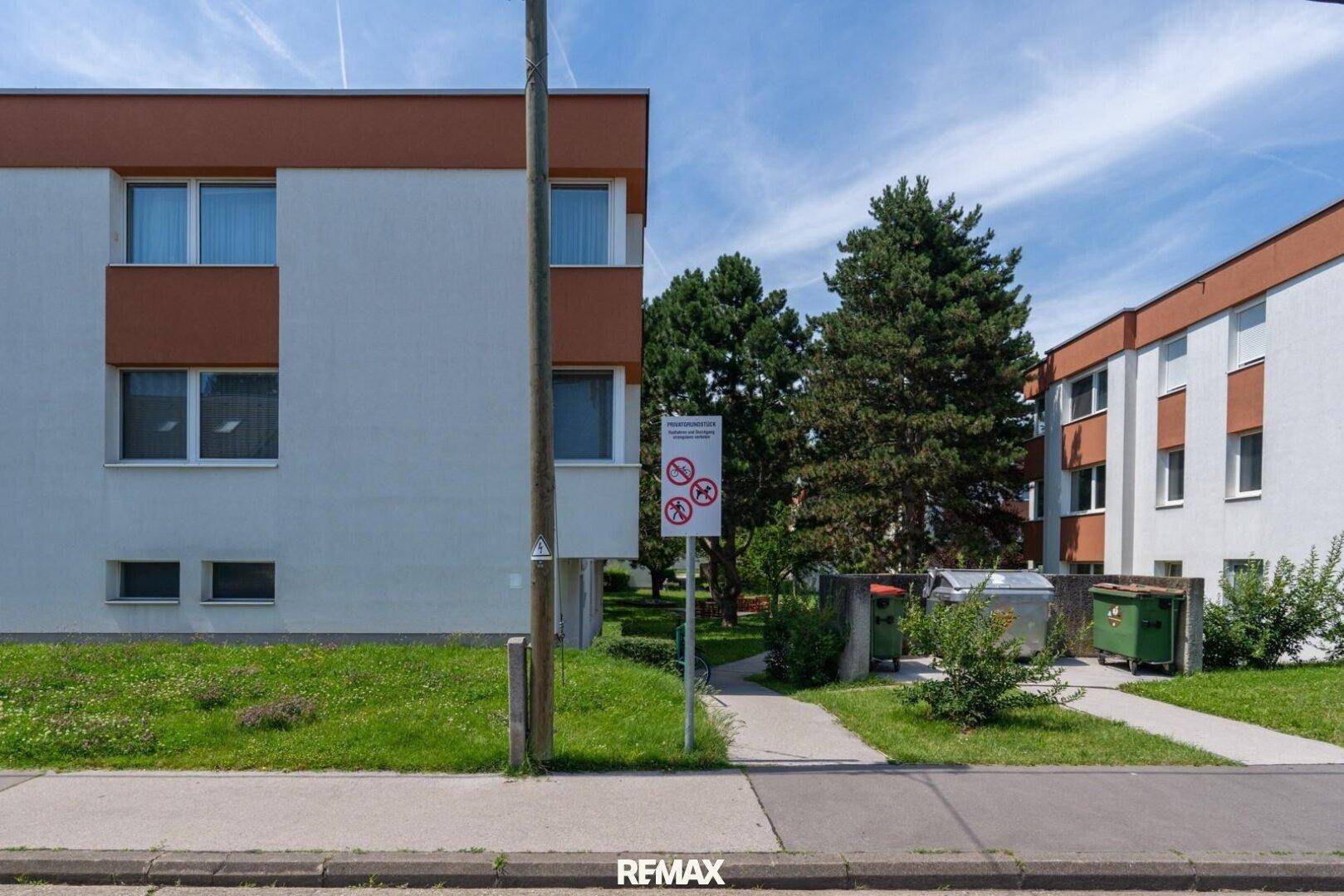 Mehrfamilienhaus mit weißer Fassade, braunen Akzenten und Mülltonnenbereich, gut in die Umgebung integriert.