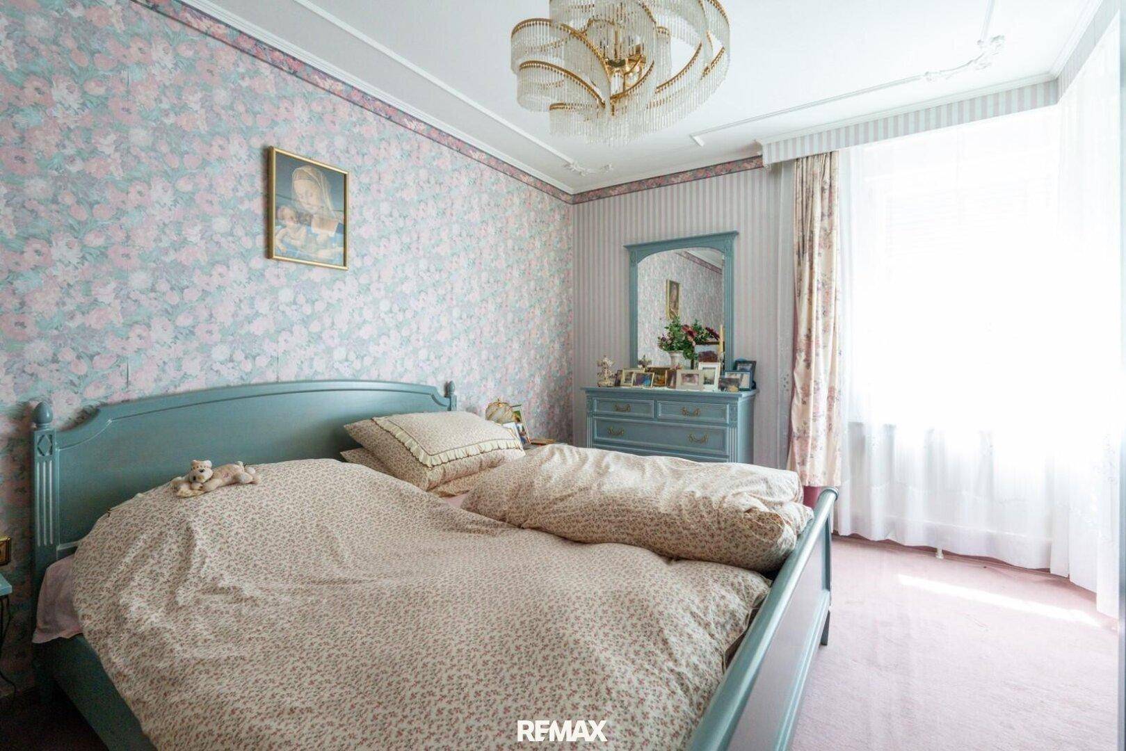 Helles Schlafzimmer mit floraler Tapete, Himmelbett und klassischem Kronleuchter, gut beleuchtet.