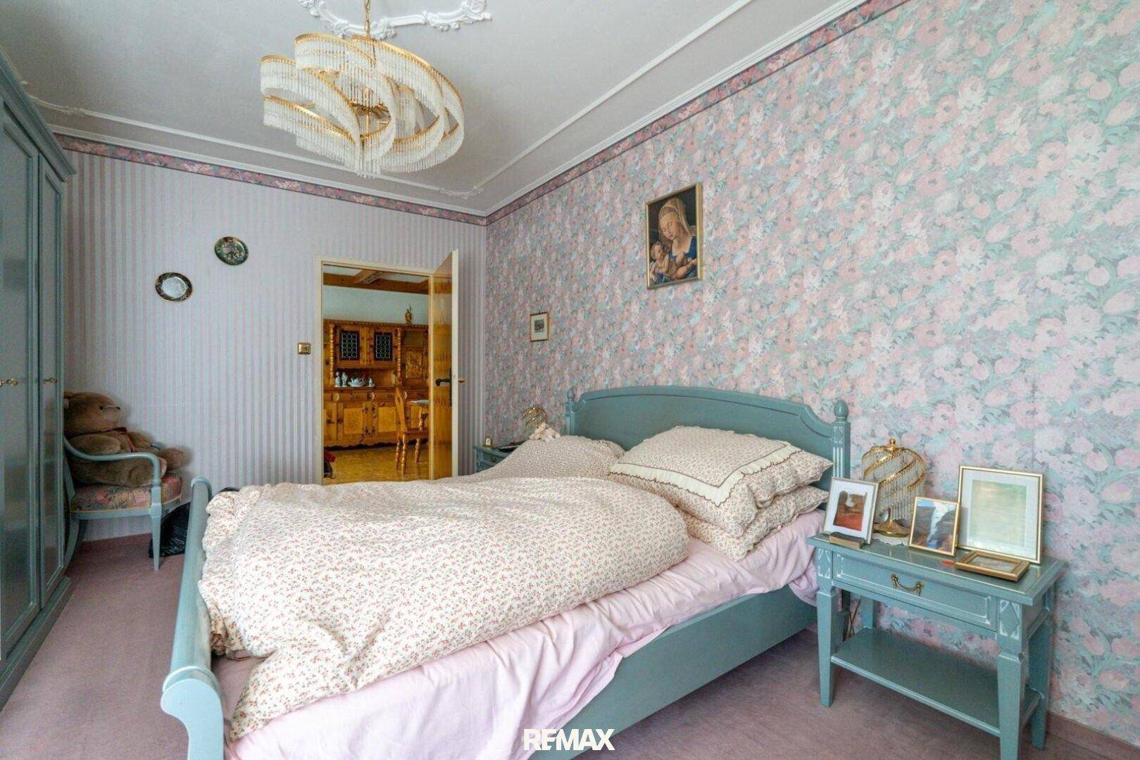 Schlafzimmer mit floraler Tapete, hellblauem Bettrahmen und Blick in den angrenzenden Flur.