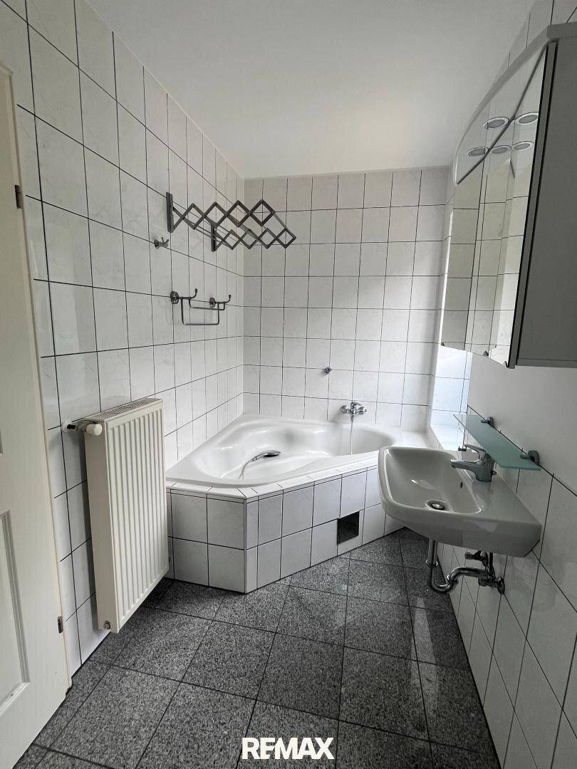 Badezimmer mit weißen Fliesen, Eckbadewanne und Waschbecken, inklusive Handtuchhalter.