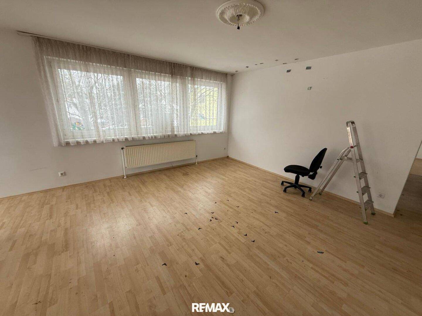 Geräumiges Wohnzimmer mit hellem Laminatboden, großen Fenstern und einem Heizkörper.