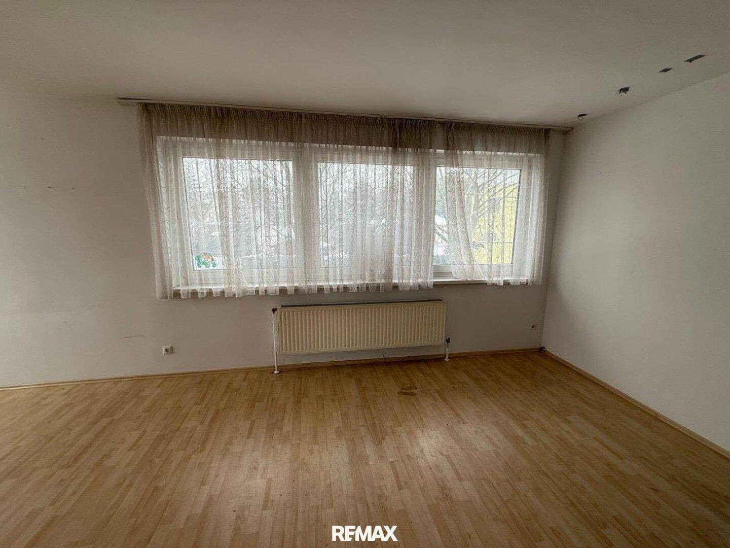 Heller Wohnbereich mit Laminatboden, großen Fenstern und einem Heizkörper.