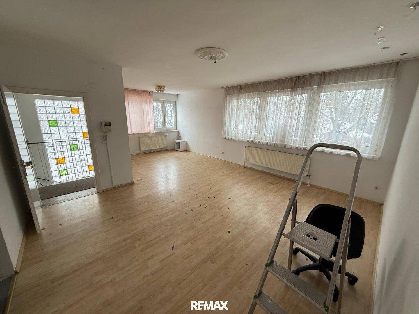 Leeres Wohnzimmer mit hellem Laminatboden, großen Fenstern und einem Heizkörper.