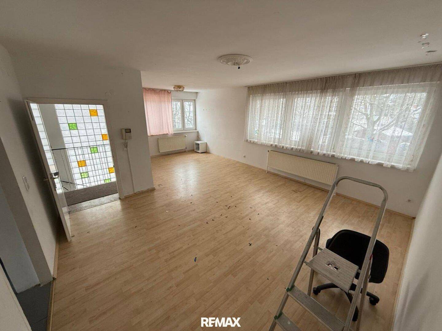 Leeres Wohnzimmer mit hellem Laminatboden, großen Fenstern und einem Heizkörper.