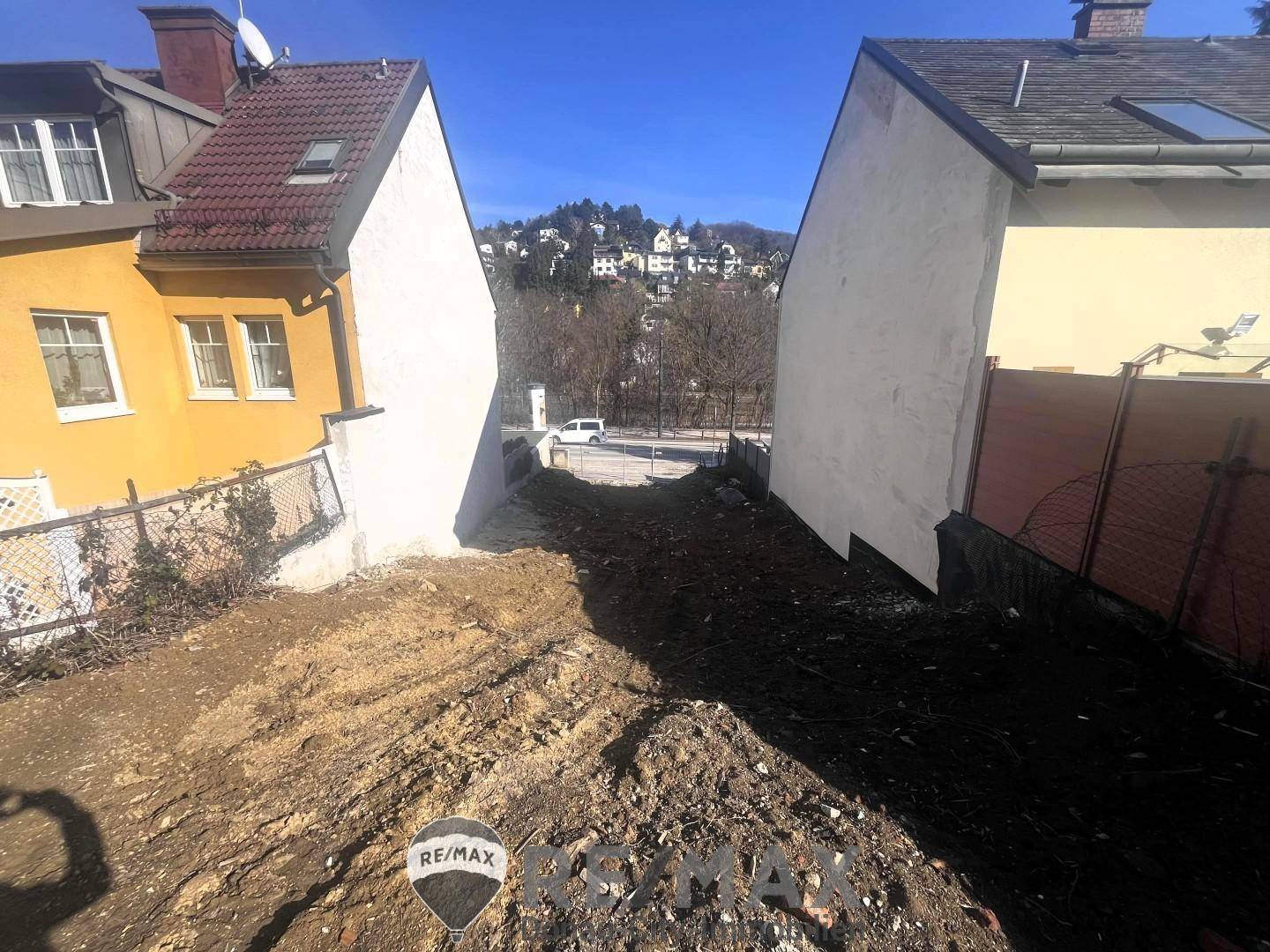 Schmaler Grundstücksbereich zwischen zwei Häusern mit Blick auf die Straße und entfernte Hügel.