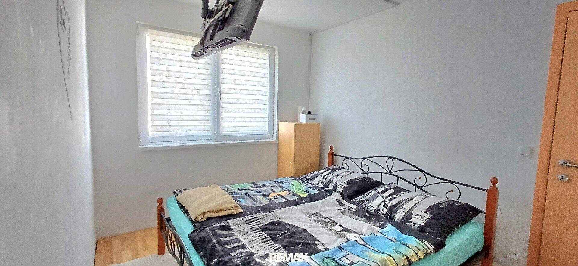 Schlafzimmer mit Doppelbett, Fenster und einem an der Decke montierten Fernseher.
