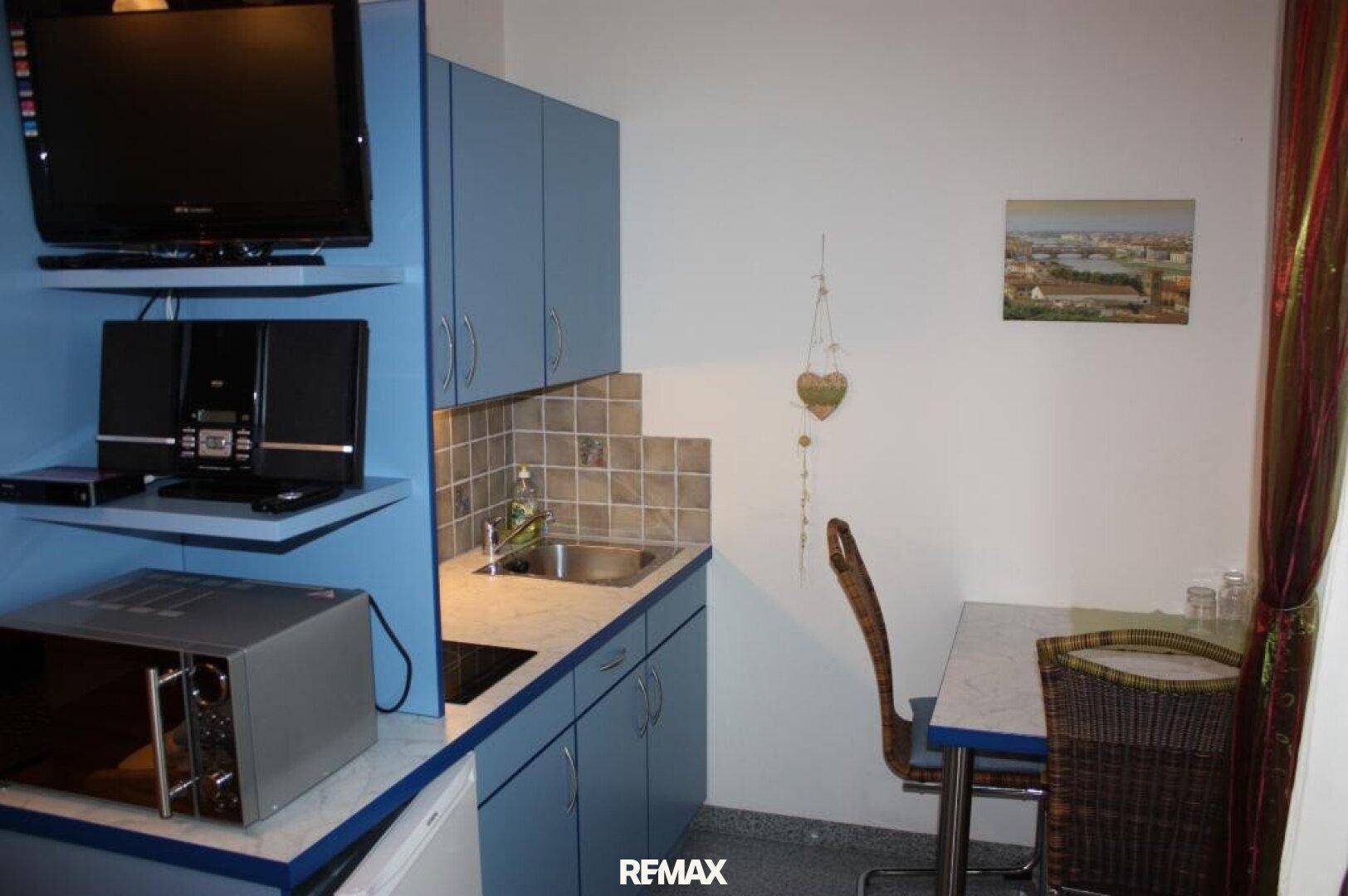Kleine Küchenzeile mit blauen Schränken, Spüle und Esstisch in einem Gästezimmer.