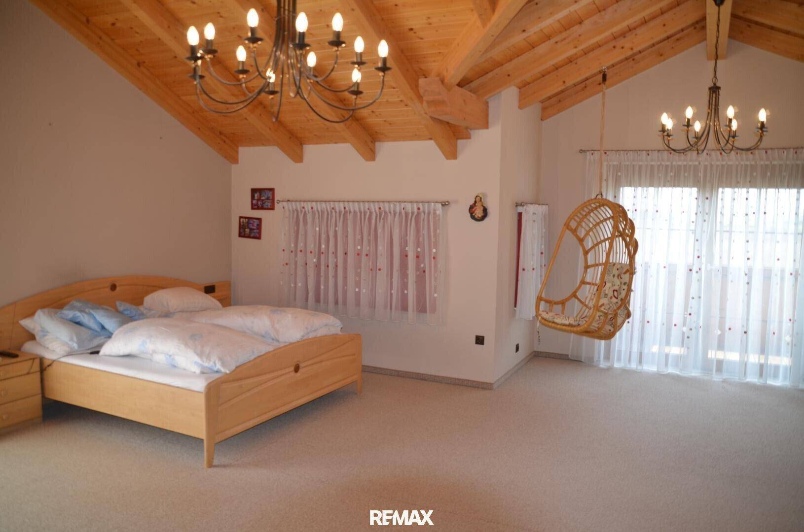 Geräumiges Schlafzimmer mit Holzbalkendecke, Teppichboden und einem Hängesessel am Fenster.