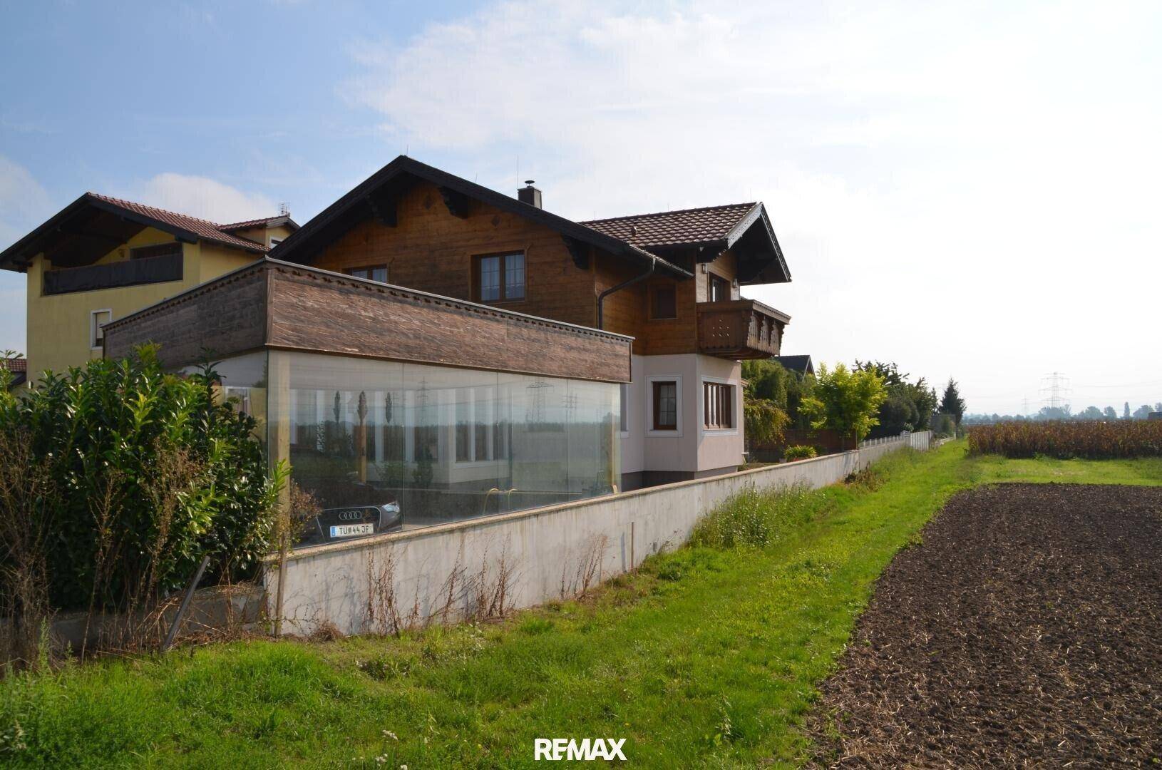 Seitenansicht eines Hauses mit Glasanbau und angrenzendem Feld, umgeben von grüner Landschaft.