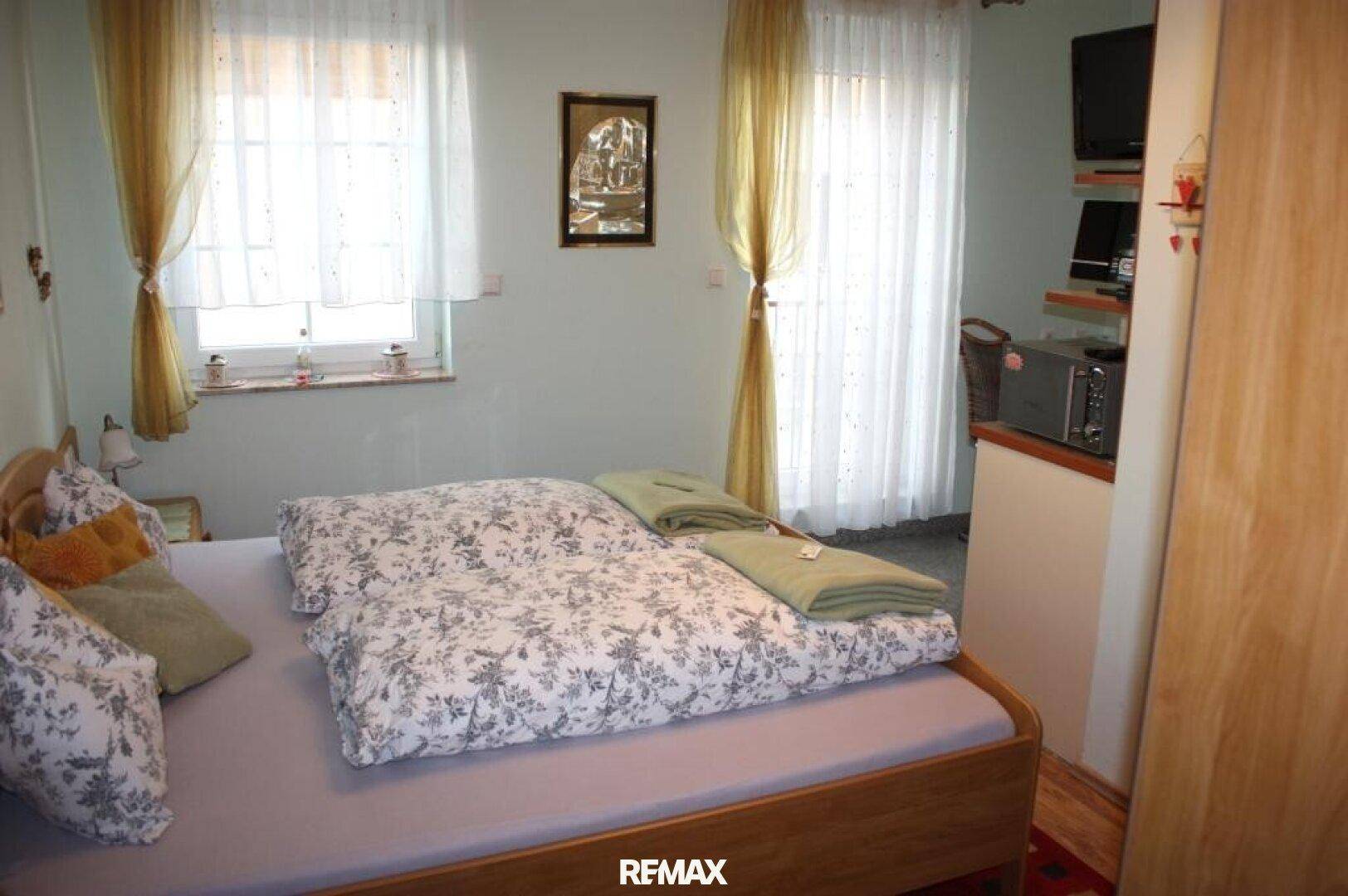 Helles Schlafzimmer mit Doppelbett, Fernseher und Fenstern mit Vorhängen.