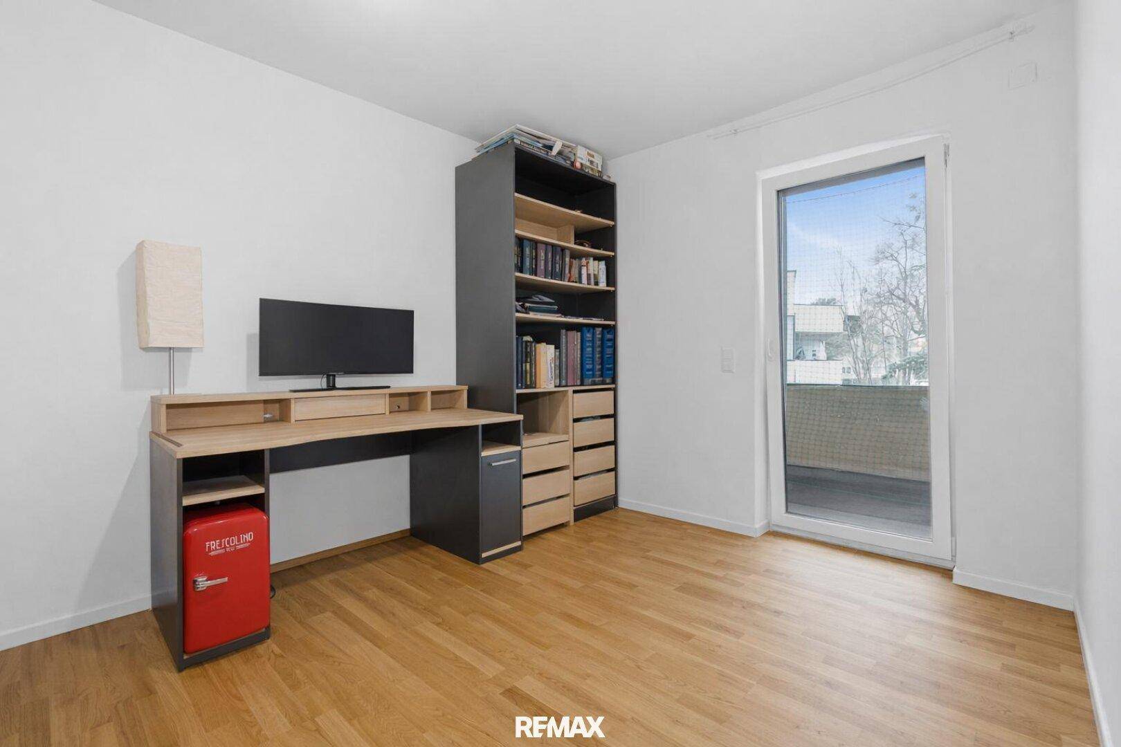 Arbeitszimmer mit Schreibtisch, Bücherregal und großem Fenster mit Zugang zum Balkon.