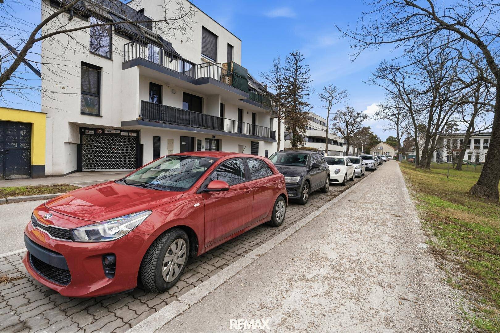 Straßenansicht mit parkenden Autos und dem modernen Wohngebäude im Hintergrund, umgeben von Bäumen.
