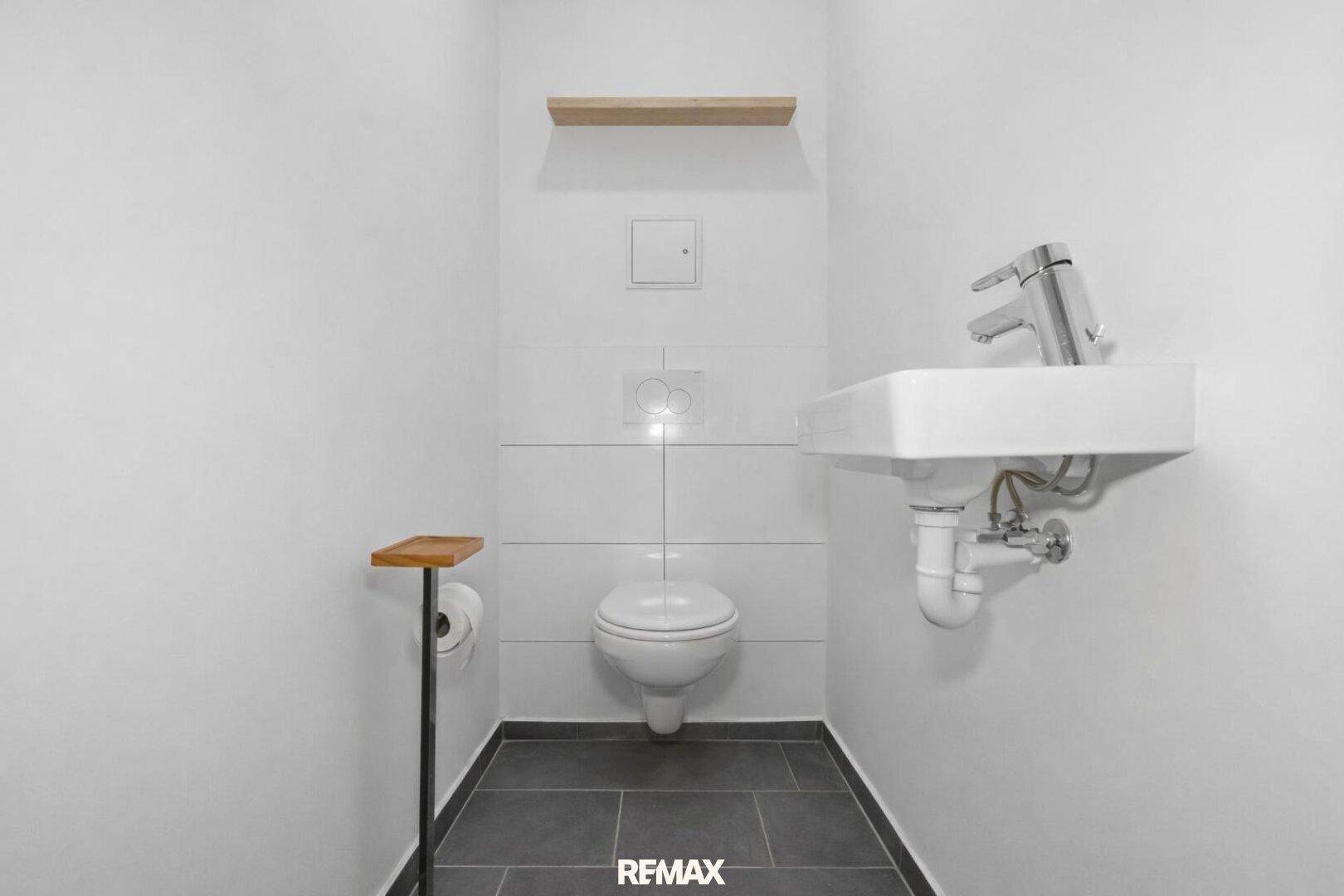 Separates Gäste-WC mit Wand-WC, kleinem Waschbecken und dunklen Bodenfliesen.