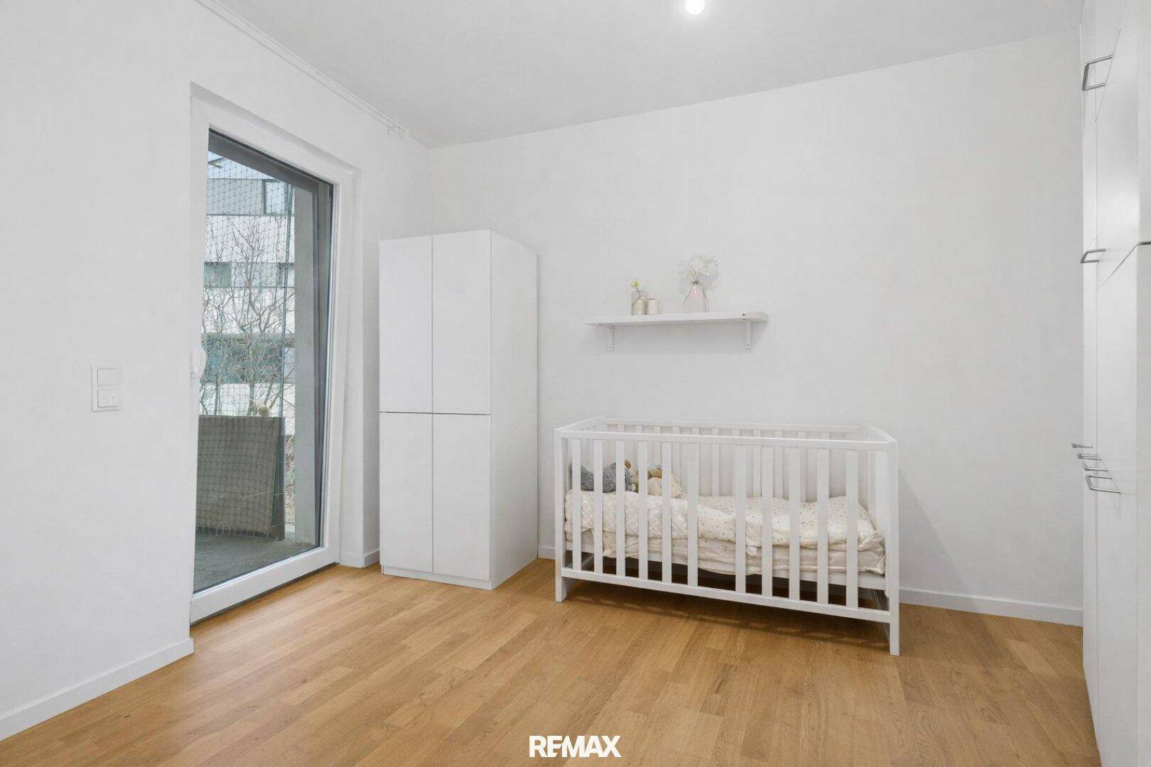 Helles Kinderzimmer mit Babybett, weißem Schrank und Zugang zu einem kleinen Balkon.