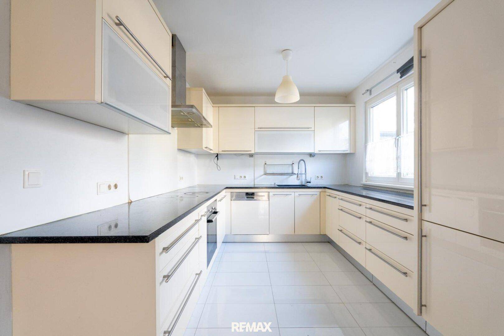 Moderne Einbauküche mit beigen Hochglanzfronten, dunkler Arbeitsplatte und Fenster.