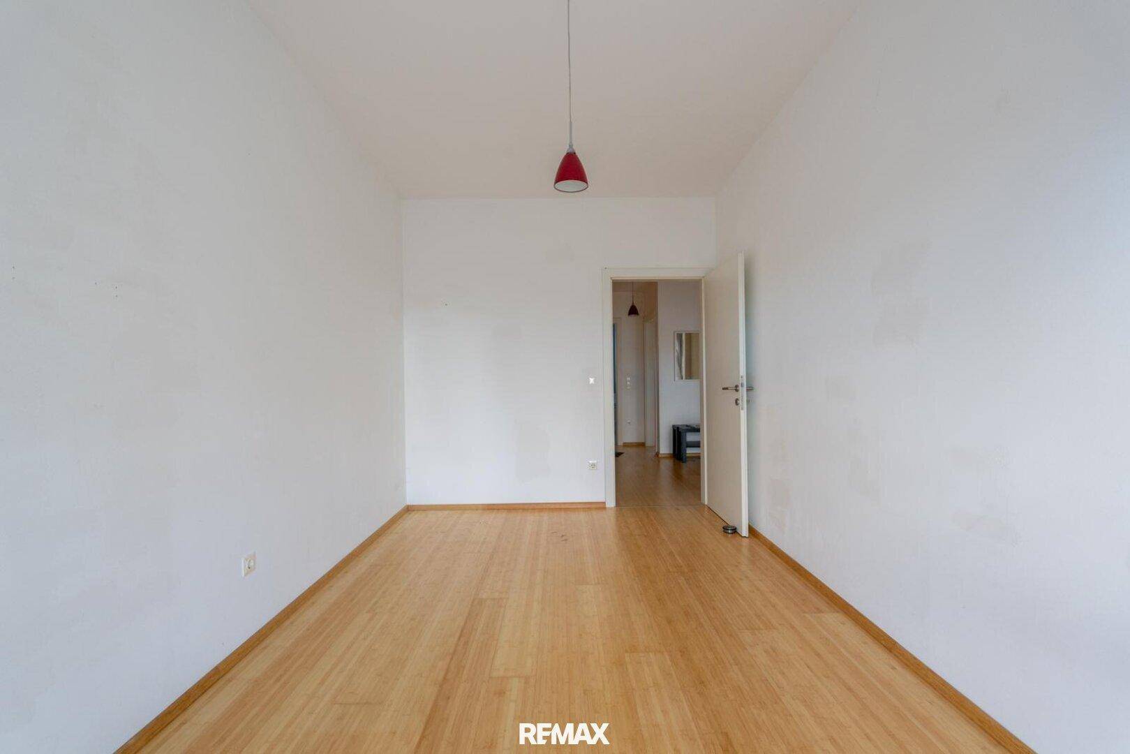 Leeres Zimmer mit hellem Holzfußboden und weiß gestrichenen Wänden, mit Tür zum Flur.
