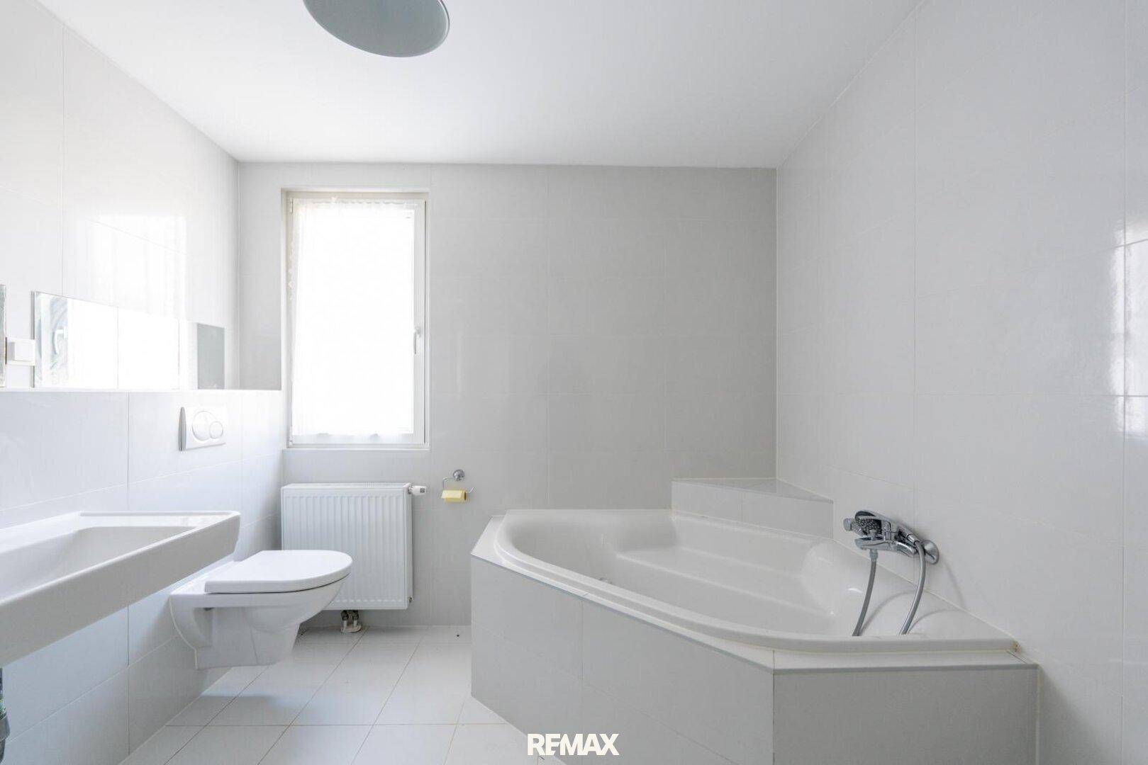 Helles Badezimmer mit weißen Fliesen, Eckbadewanne und Fenster für natürliche Belüftung.