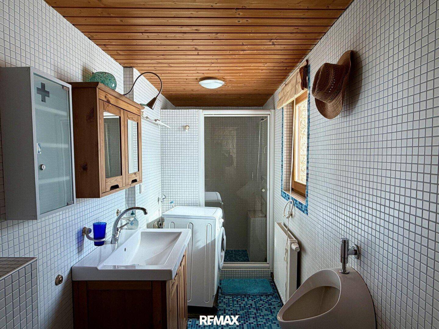 Kleines Badezimmer mit Dusche, Urinal, Waschmaschine und weißen Mosaikfliesen.