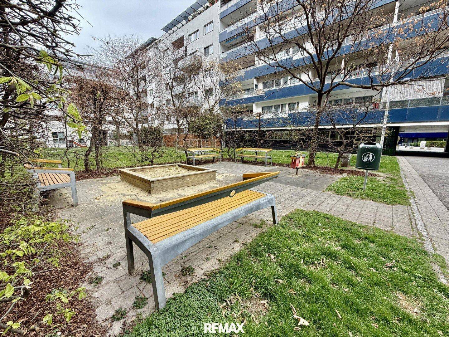 Gemeinschaftlicher Gartenbereich mit Sandkasten und Bänken, umgeben von Bäumen.