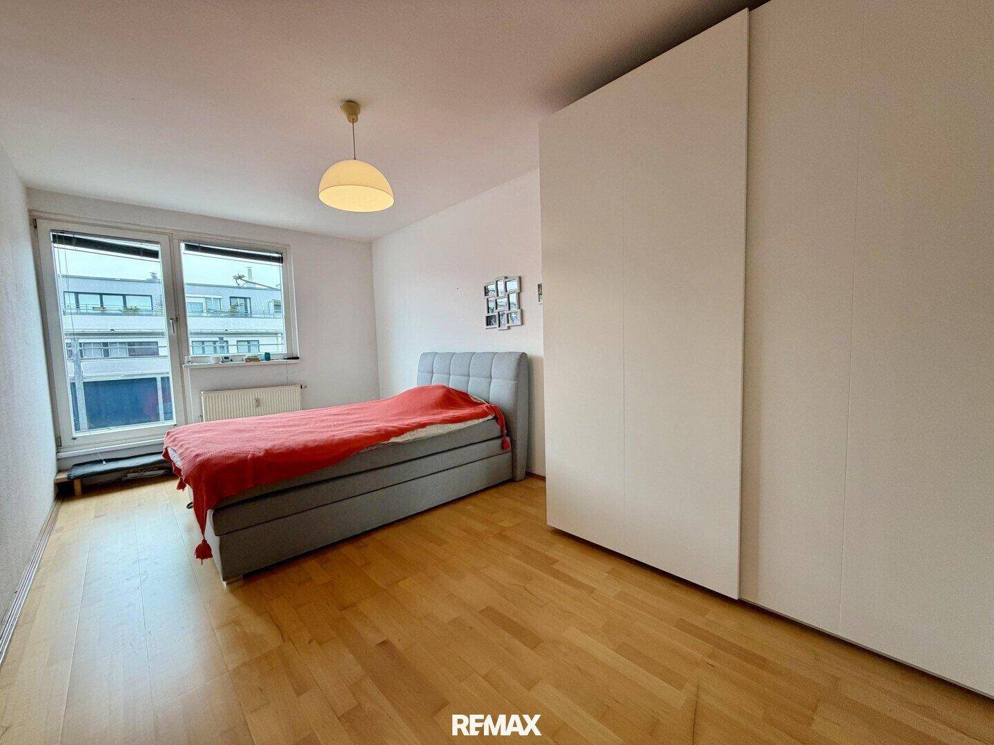 Helles Schlafzimmer mit Holzboden, großem Fenster und einem modernen Bett mit roter Decke.