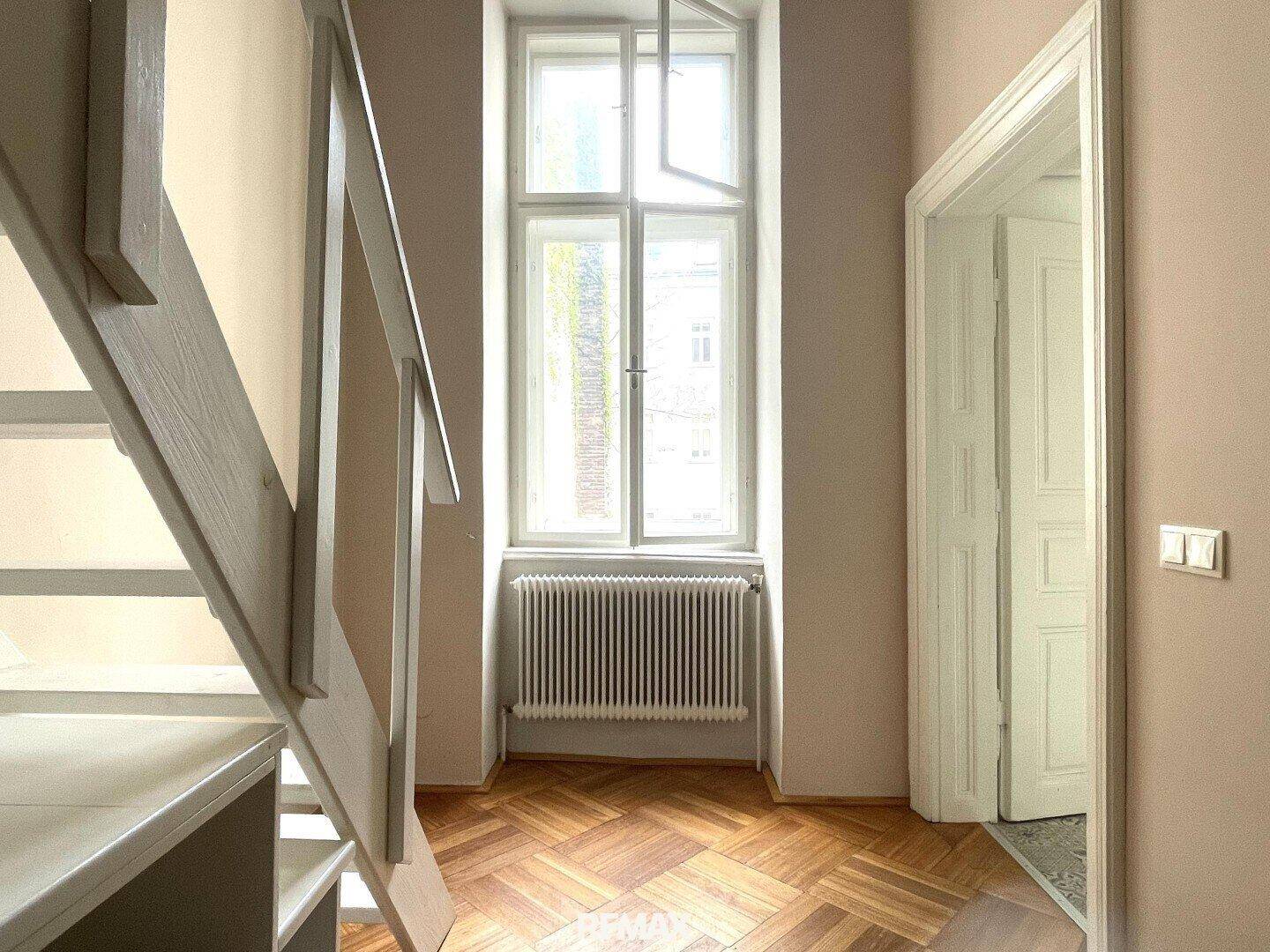 Heller Flur mit Treppe, hohem Fenster und Fischgrätparkettboden, der zum Obergeschoss führt.