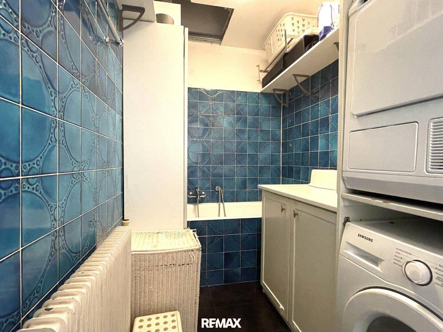 Praktischer Waschraum mit blauen Fliesen, Badewanne, Waschmaschine und Trockner.