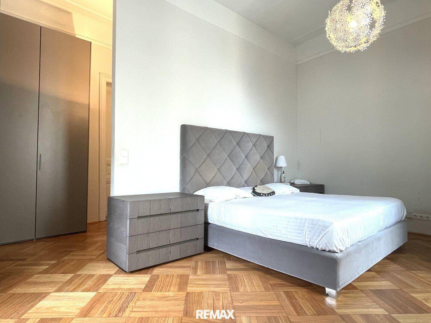 Schlafzimmer mit grauem Polsterbett, passenden Nachttischen und Kleiderschrank, Parkettboden.
