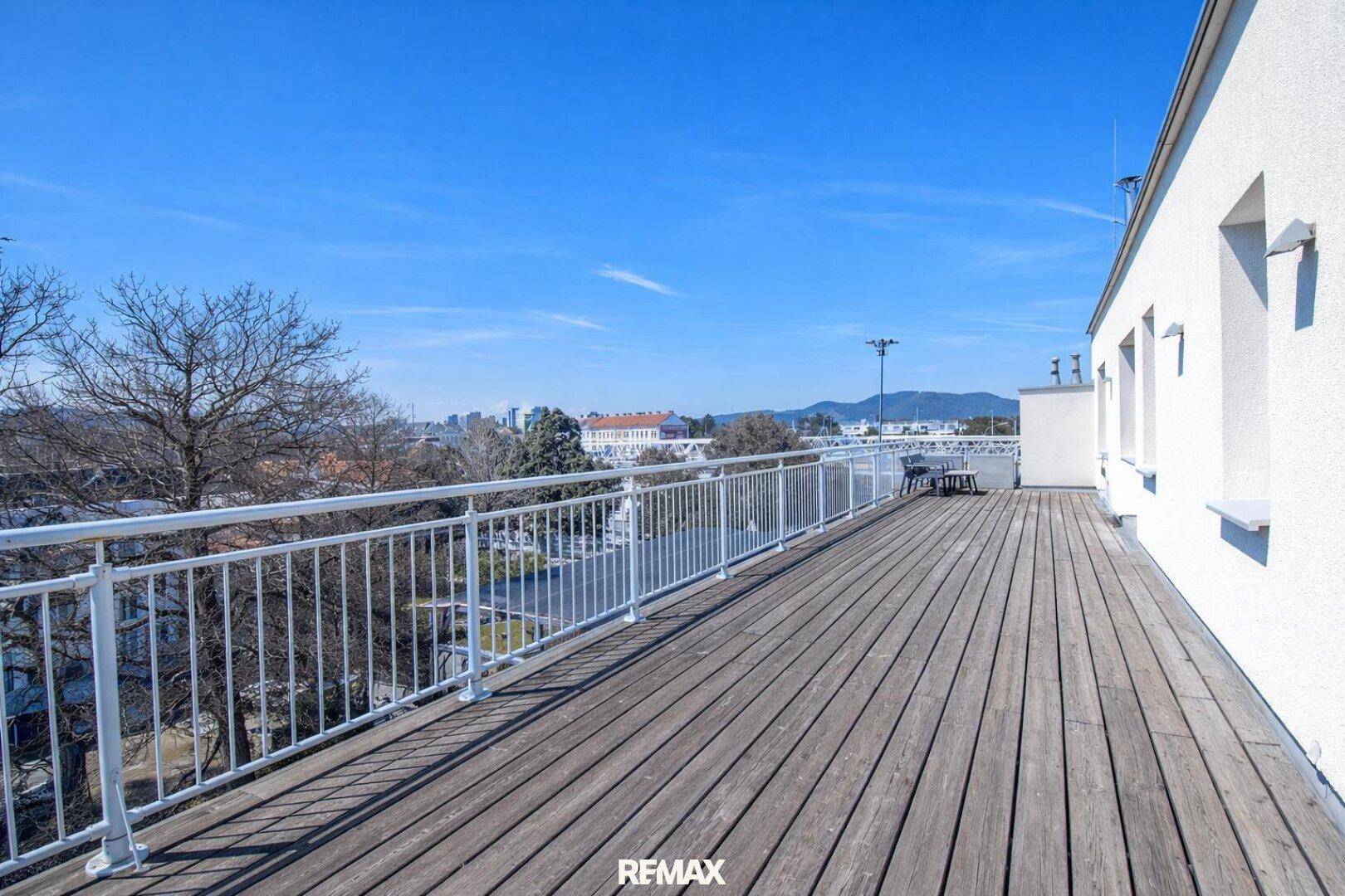 Lange, sonnige Terrasse mit Holzboden und weißem Geländer, bietet einen weiten Blick über die Stadt.