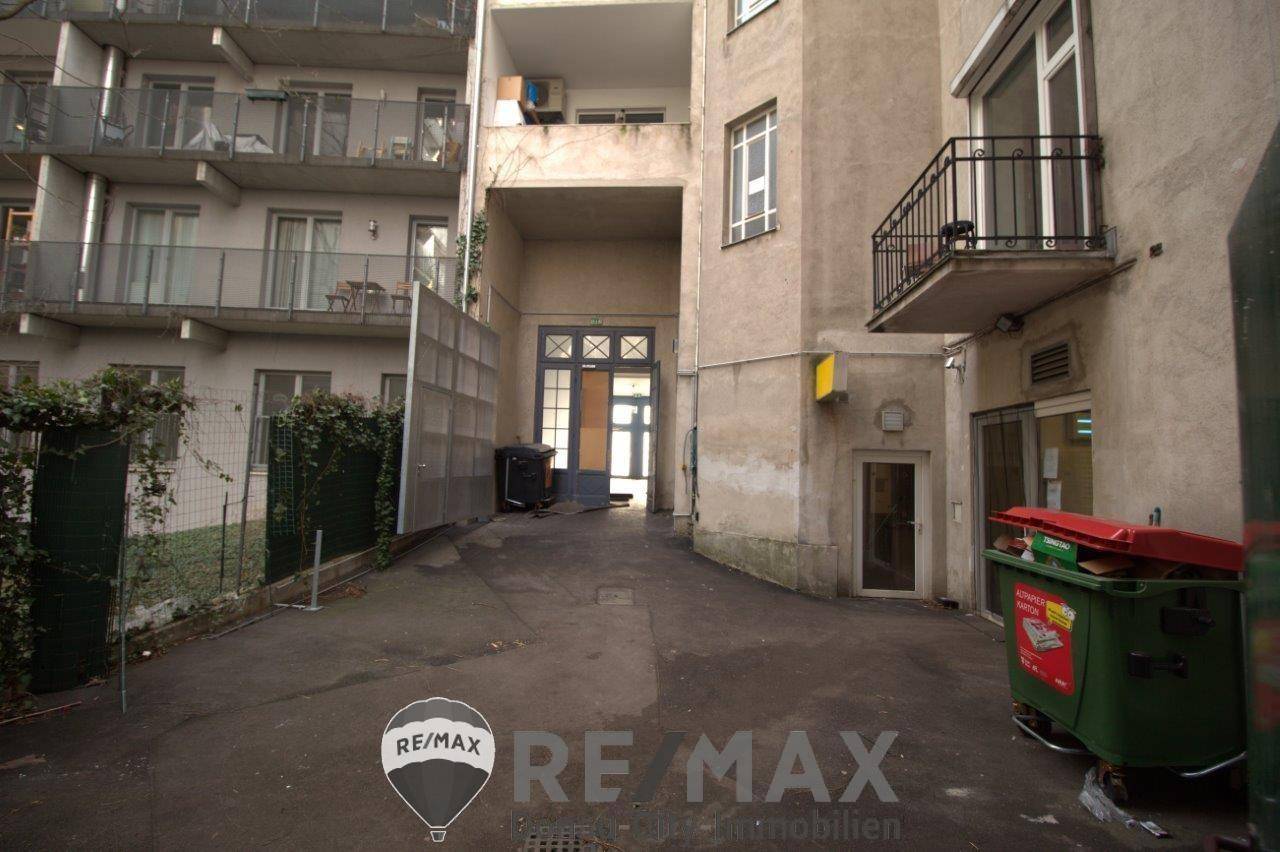 Eingangsbereich eines Gebäudes mit Innenhof, umgeben von weiteren Gebäudeteilen und sichtbaren Mülltonnen.