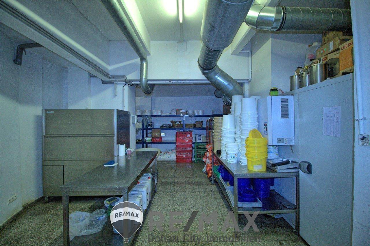 Funktionaler Lagerraum oder Vorbereitungsbereich mit Metallregalen, Tischen und sichtbaren Lüftungsrohren an der Decke.
