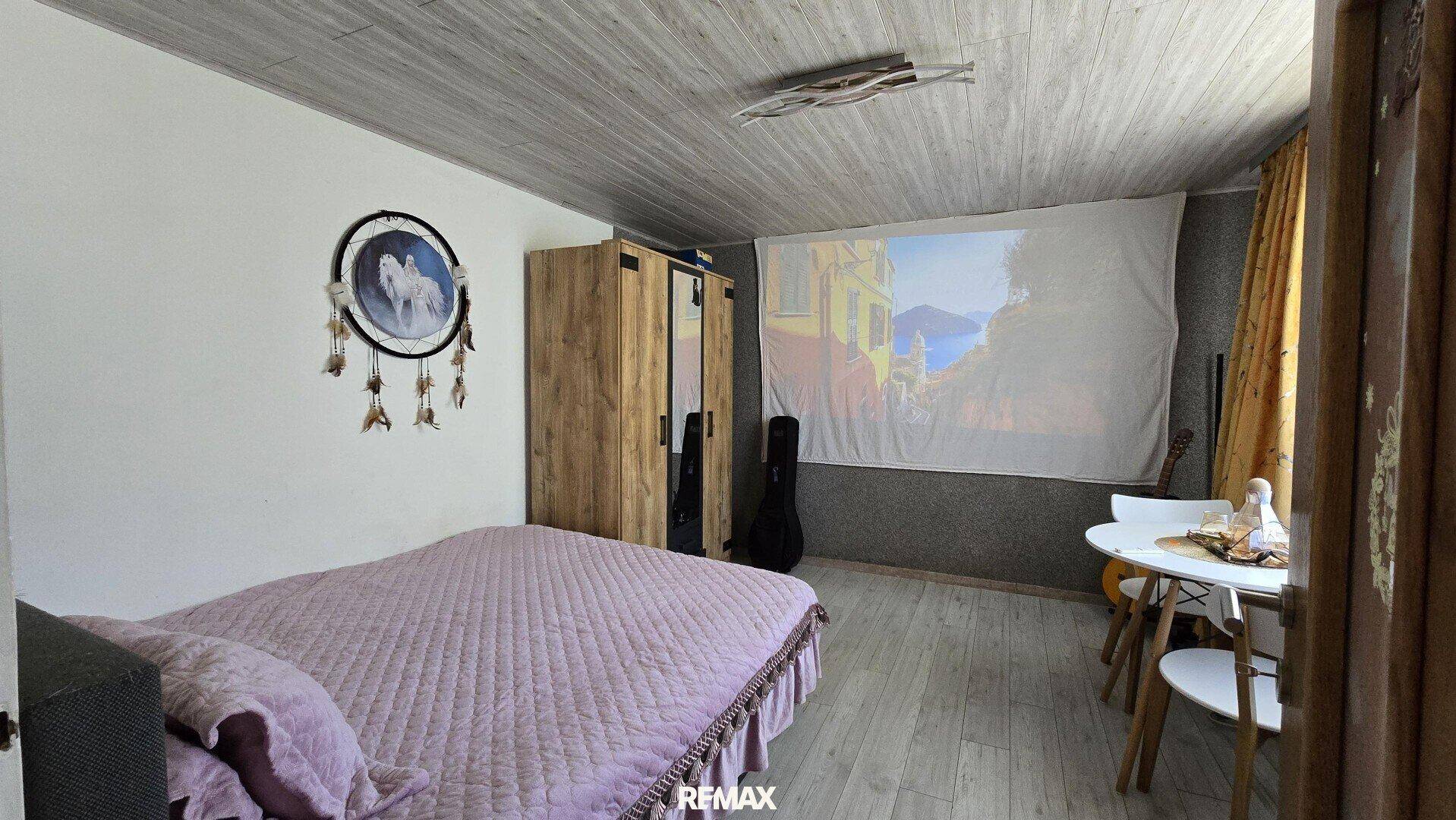 Schlafzimmer mit Doppelbett, Holzschrank und Projektionsfläche an der Wand.
