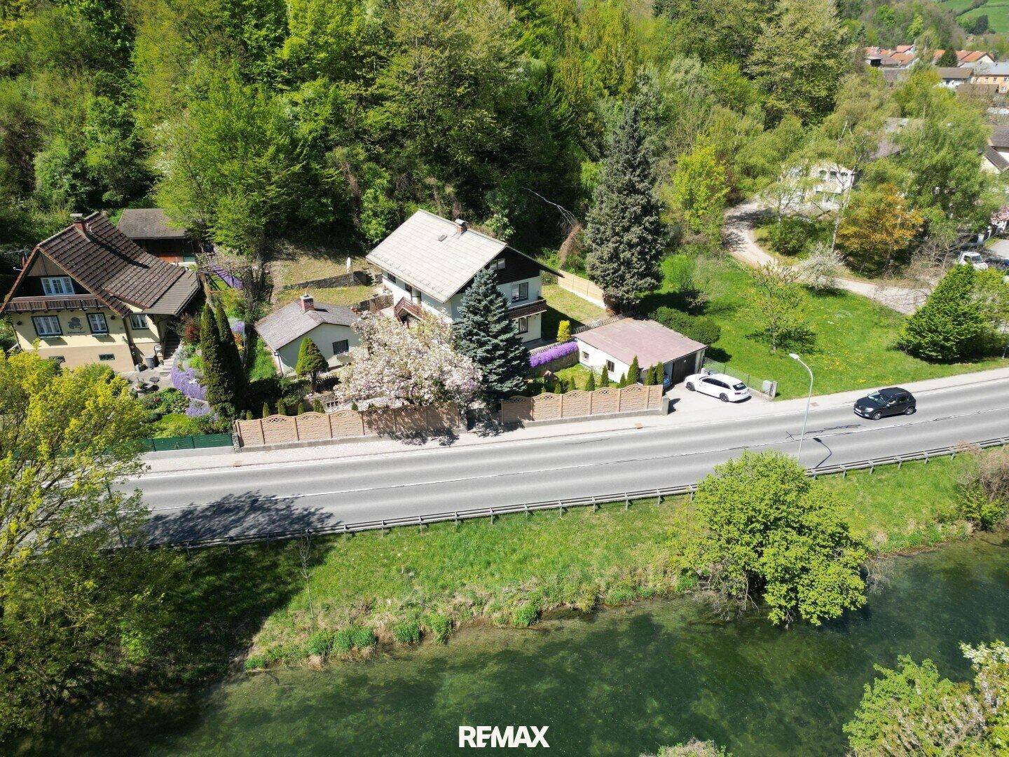 Weitläufige Luftaufnahme des Anwesens mit Haus, Garten, Straße und einem Fluss im Vordergrund.