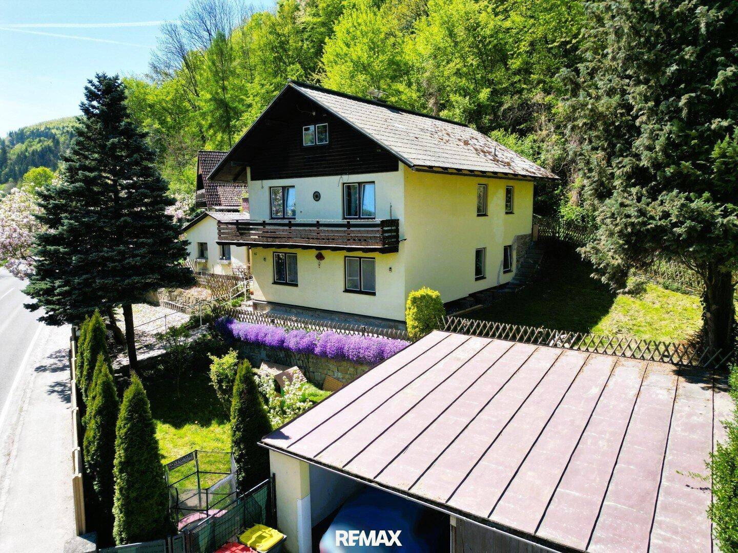 Luftaufnahme eines Hauses mit Garten, Balkon und Garage, umgeben von grünen Hügeln und Bäumen.