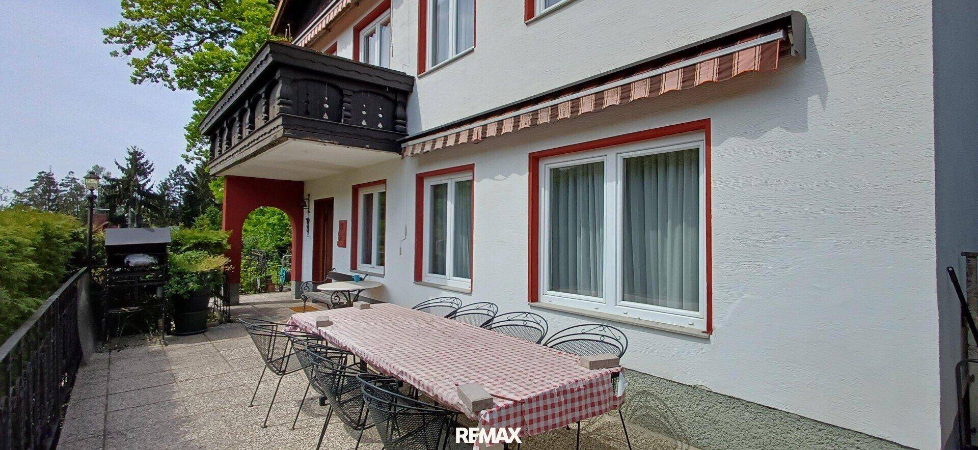 Hausfassade mit großer gepflasterter Terrasse, Markise und einem kleinen Holzbalkon.