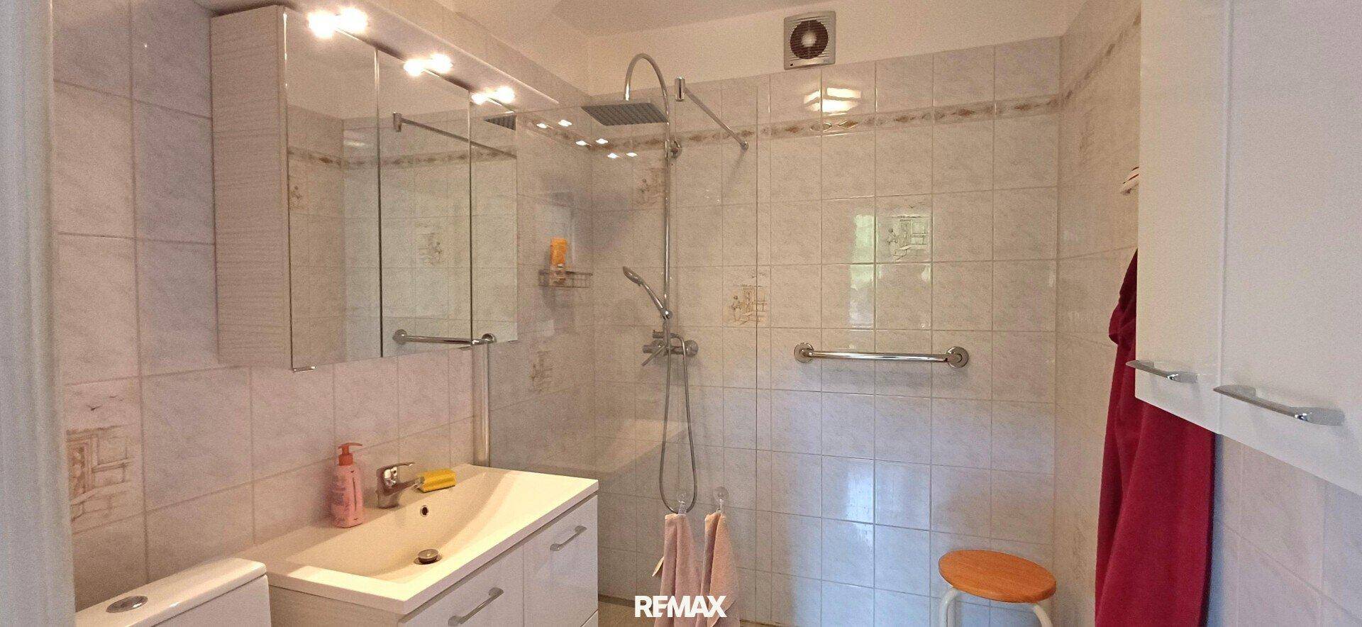 Helles Badezimmer mit weißen Fliesen, Dusche und einem Spiegelschrank über dem Waschbecken.