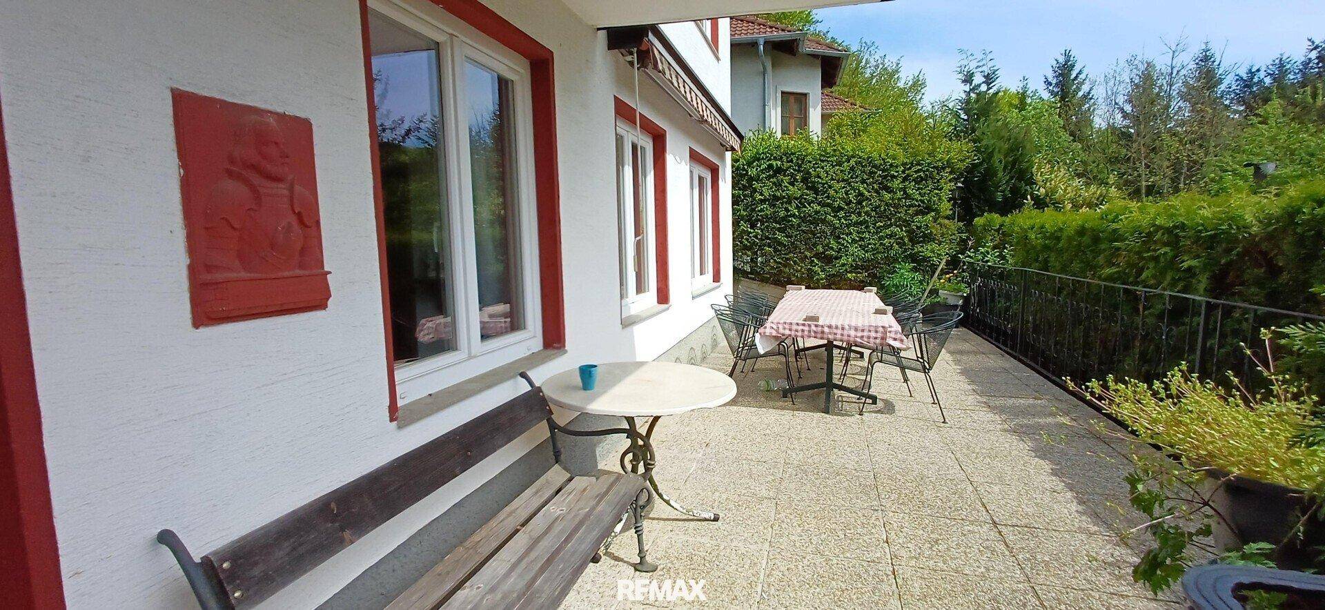 Gemütliche Terrasse mit Esstisch und Stühlen, umgeben von Hecken und mit Blick ins Grüne.