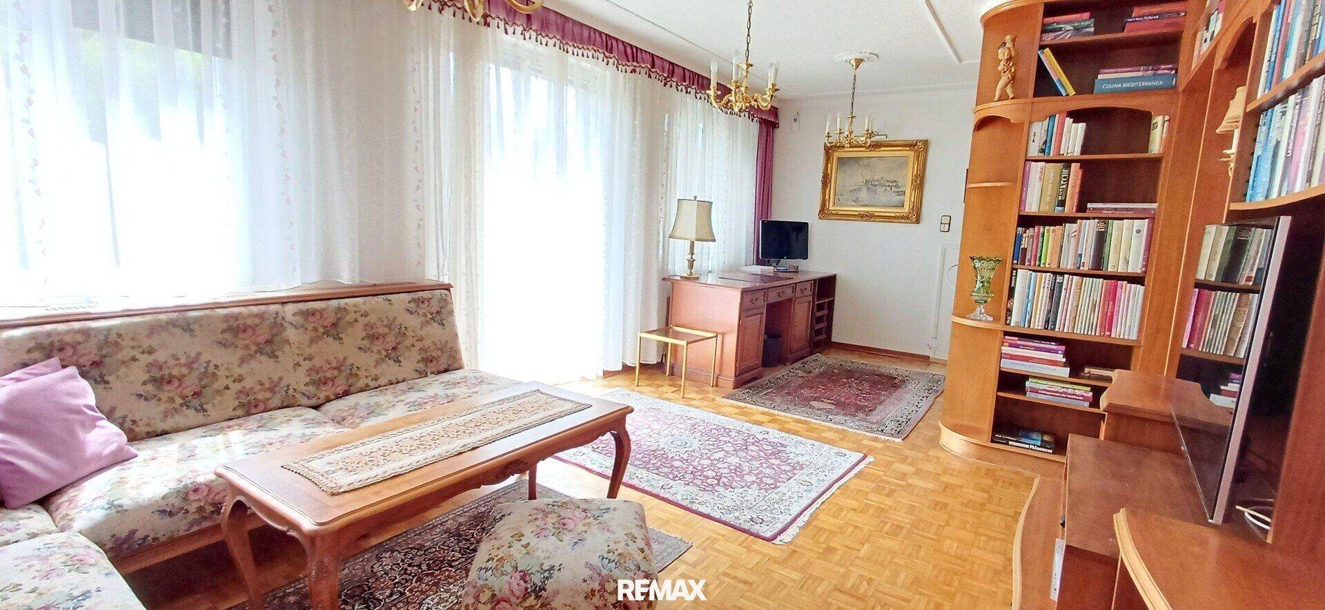 Heller Wohnbereich mit großem Fenster, Sofa, Schreibtisch und Bücherregalen.