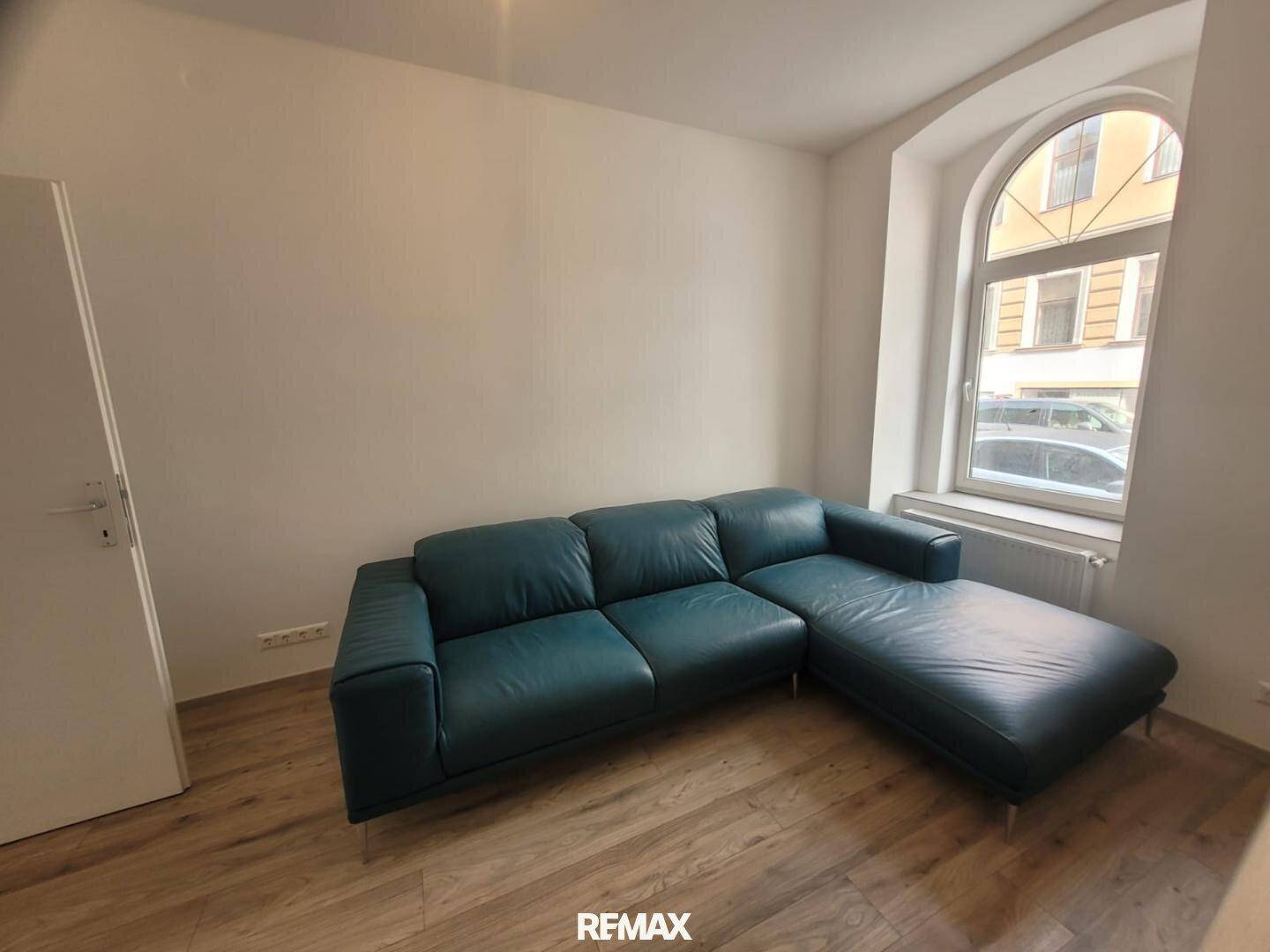 Gemütliches Wohnzimmer mit einem blauen Ledersofa und Laminatboden, hell durch ein Rundbogenfenster.