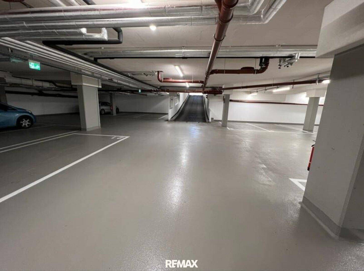 Helle Tiefgarage mit Zufahrtsrampe, sichtbaren Rohren und Brandschutzvorrichtungen.