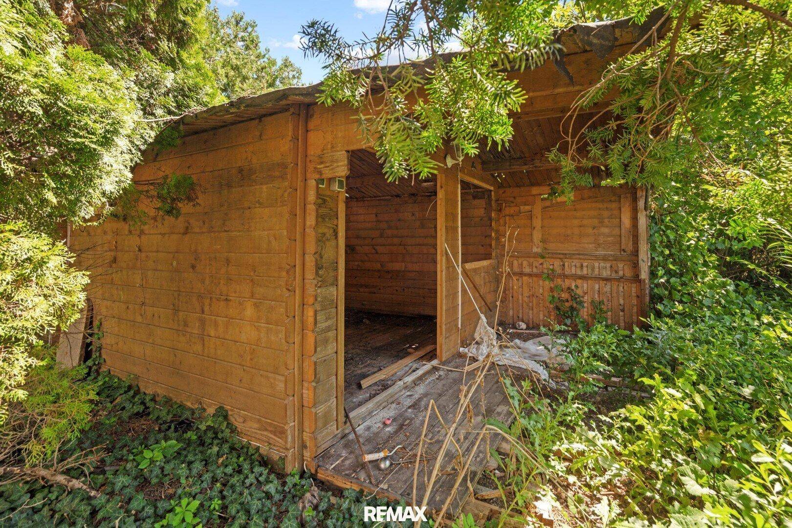 Rustikaler Holzschuppen im Gartenbereich, umgeben von Bäumen und Sträuchern.