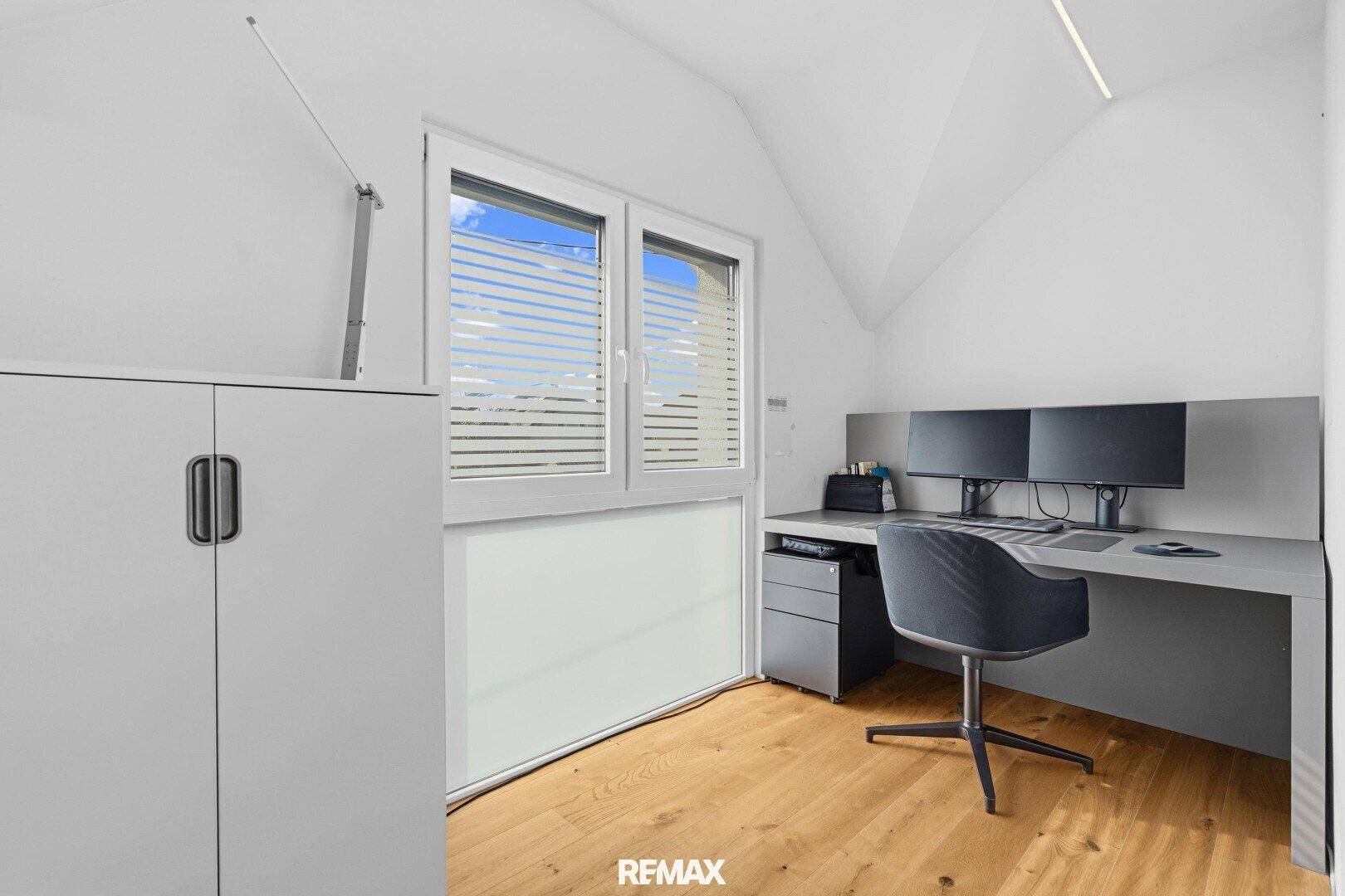 Helles Arbeitszimmer mit Schreibtisch, zwei Monitoren, Holzboden und Dachschräge.