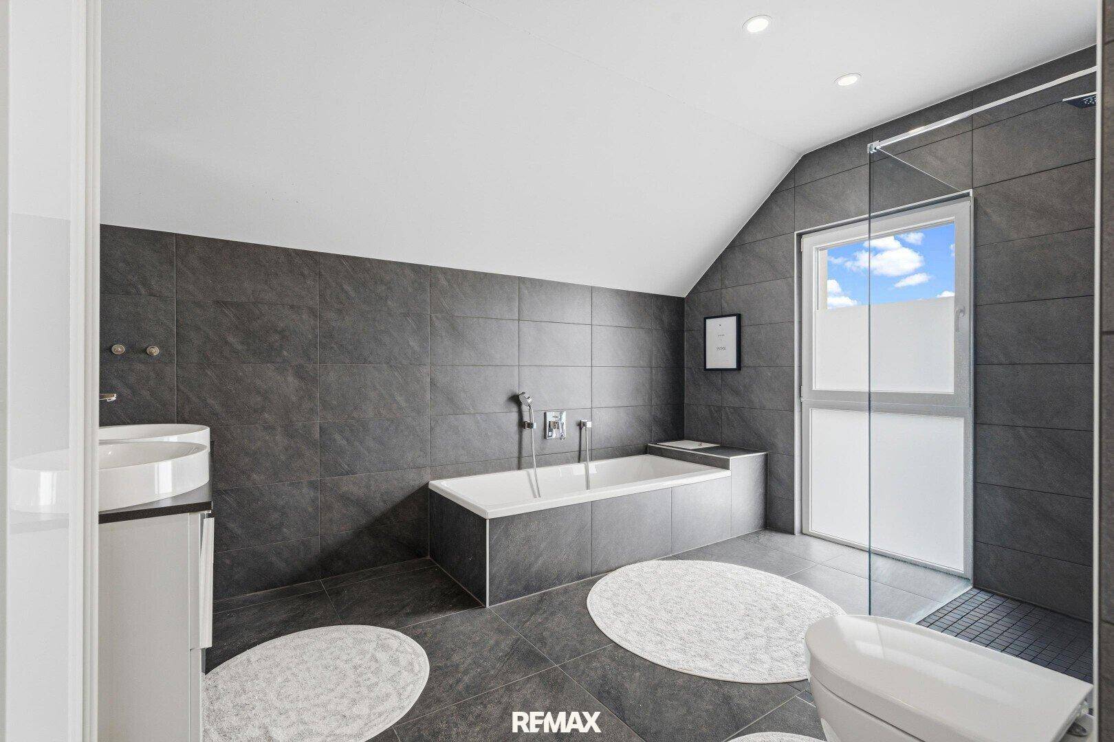 Geräumiges Badezimmer mit dunklen Fliesen, Badewanne, Dusche und Dachschräge.