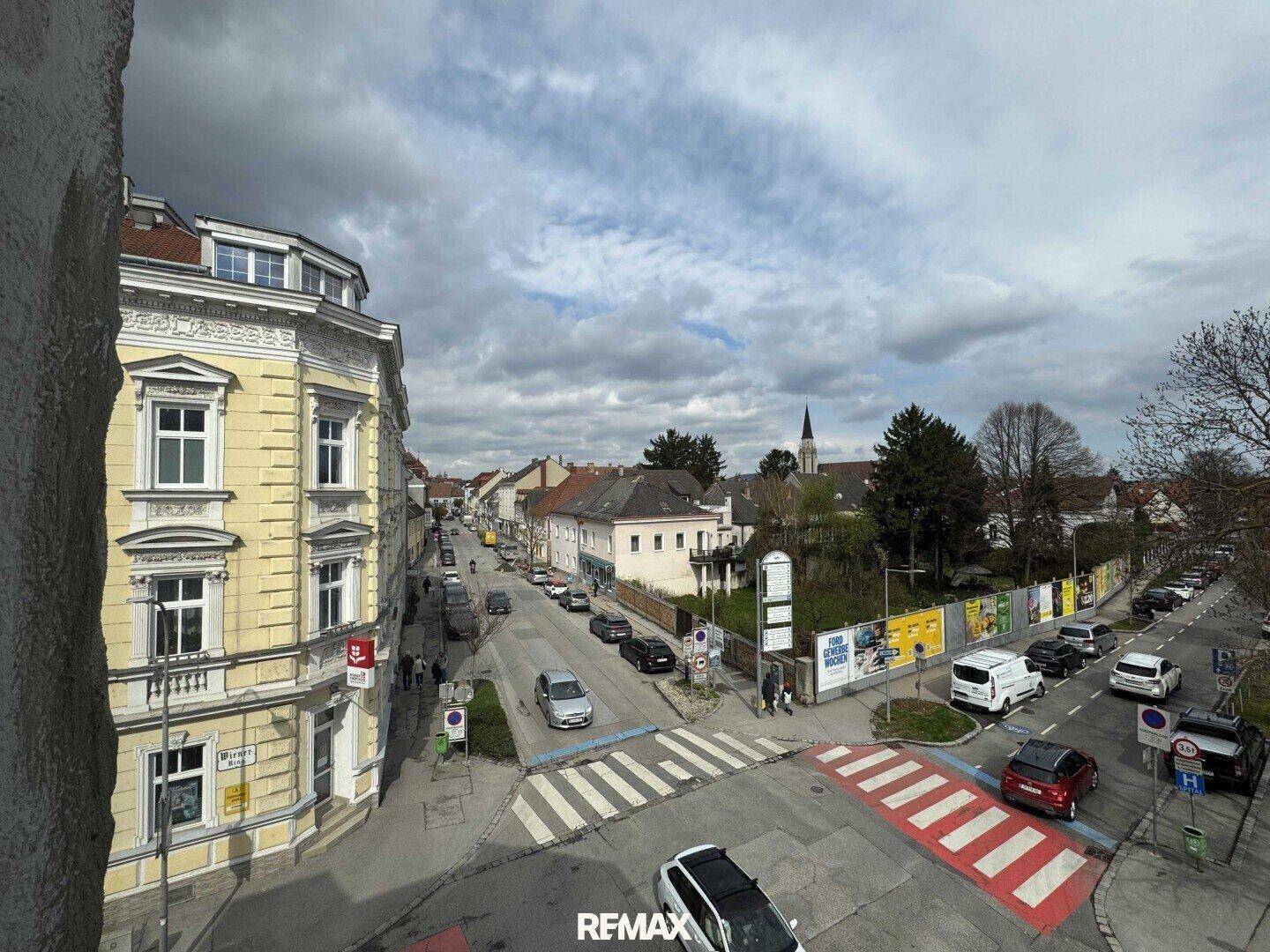 Blick auf eine belebte Stadtstraße mit parkenden Autos und Gebäuden unter bewölktem Himmel.