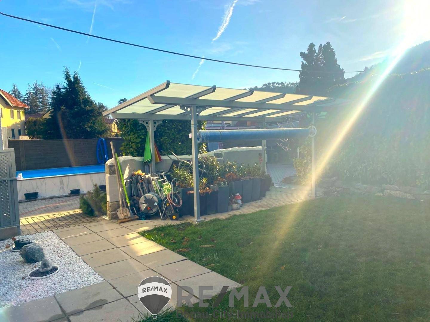 Gartenbereich mit Carport, Rasenfläche und verschiedenen Pflanzen unter strahlendem Sonnenschein.