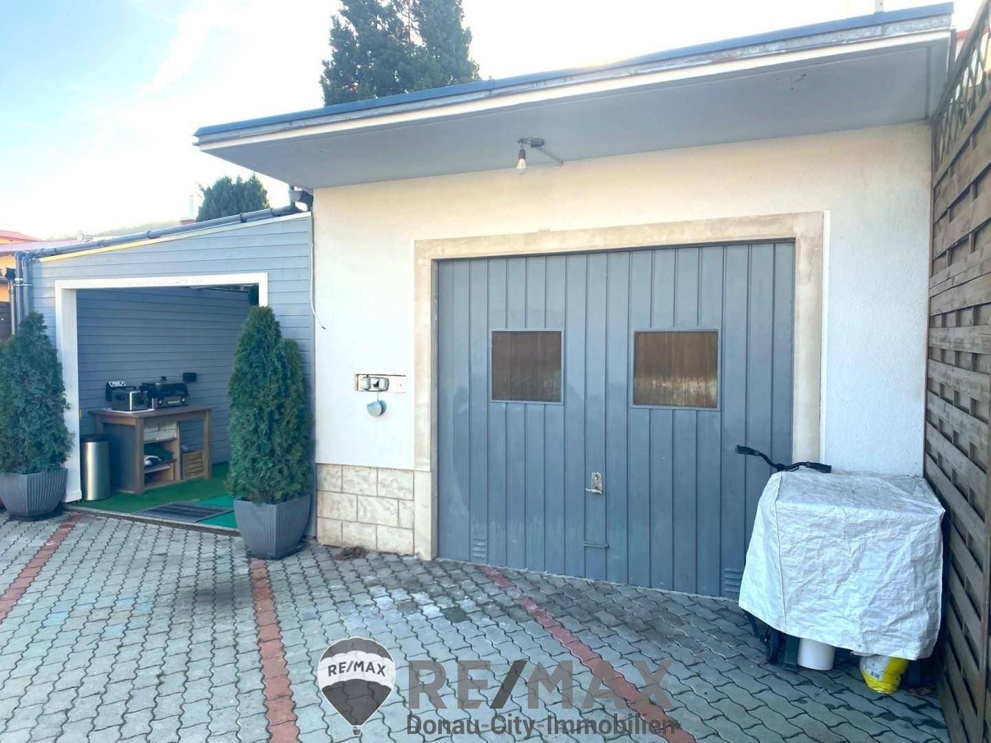 Garage mit Holztor und gepflasterter Zufahrt, daneben ein weiterer überdachter Bereich.
